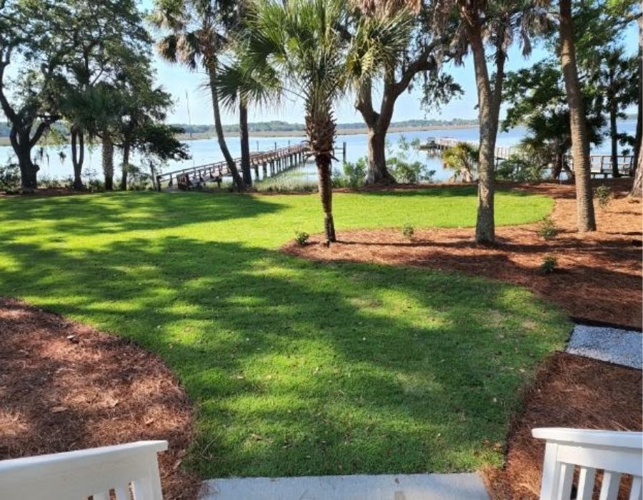 Waterfront backyard professionally landscaped by Greenspae HHI in the Lowcountry, featuring lush green grass, palm trees, pine straw beds, and a private dock overlooking scenic coastal waters