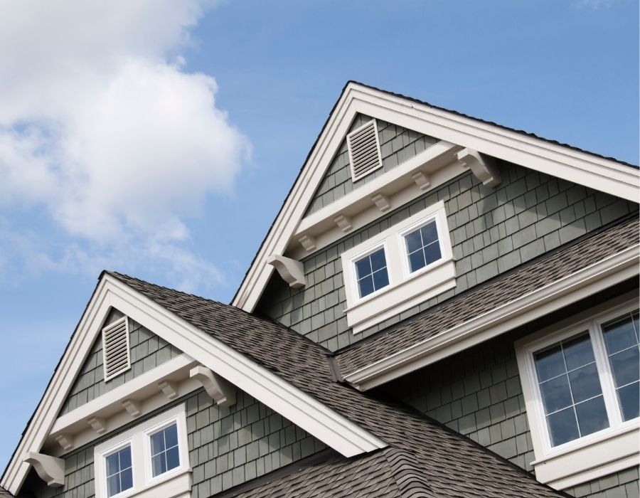 Close-up view of a Lowcountry home featuring gray shake siding, architectural roof shingles, white trim, and gable vents, highlighting coastal-inspired exterior design and durable roofing materials