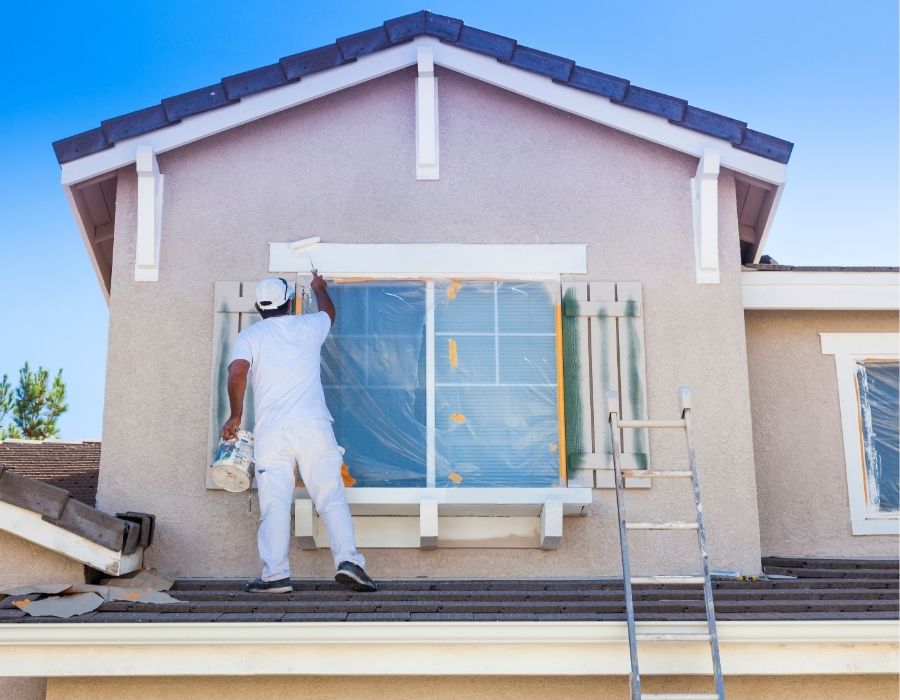 Professional painter from The Painting Pros in the Lowcountry applying fresh white trim paint to a second-story window frame on a stucco home, demonstrating expert exterior painting services and attention to detail